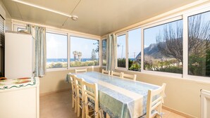 A dining area with a table and chairs, large windows with a view of the sea and mountains, and a refrigerator.