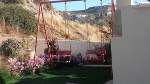 A red swing set in a backyard with a grassy lawn, potted plants, and a hillside in the background. - Apartment 'No 3' with Sea View, Wi-Fi and Air Conditioning (Kos)
