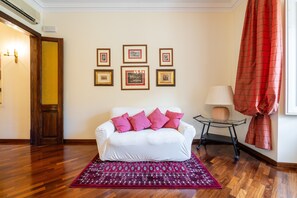 A white sofa with pink cushions, a patterned rug, framed pictures on the wall, and a red curtain. - Apartment 'Amita' with Wi-Fi and Air Conditioning (Cagliari)