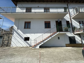 A two-story building with a staircase leading to the upper floor, a stone wall to the left, and a balcony with metal railings.