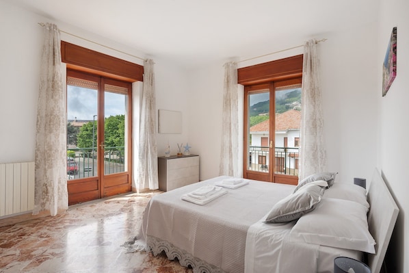 A bedroom with a bed, curtains, a dresser, and a view of buildings and trees.