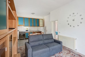 A living room with a grey sofa, a kitchen in the background, and a wall clock.