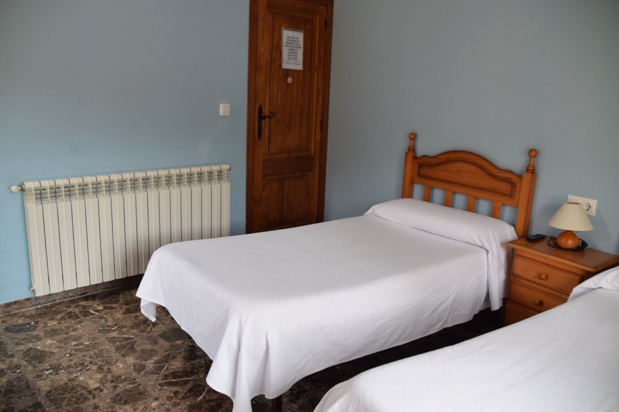 Una habitación con dos camas individuales, una puerta de madera, un radiador y una mesita de noche con una lámpara.