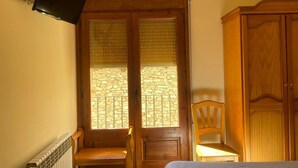 A room with a bed, a wooden wardrobe, a wooden chair, a bench, a television mounted on the wall, and a window with blinds. - Room 'Twin Con Balcón' with Balcony and Wi-Fi (Villarluengo)