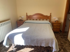 Une chambre à coucher avec une tête de lit en bois, un lit recouvert d’une courtepointe blanche, deux tables de chevet avec des lampes, un radiateur et un sol en marbre.