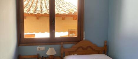 A bedroom with a bed, a bedside table with a lamp, a nightstand, and a window with a view of a tiled roof.