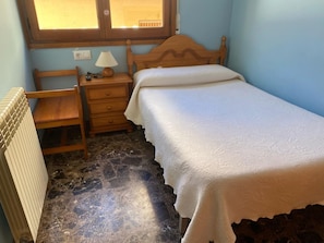 A small bedroom with a single bed, a wooden nightstand, a chair, a radiator, and a window.