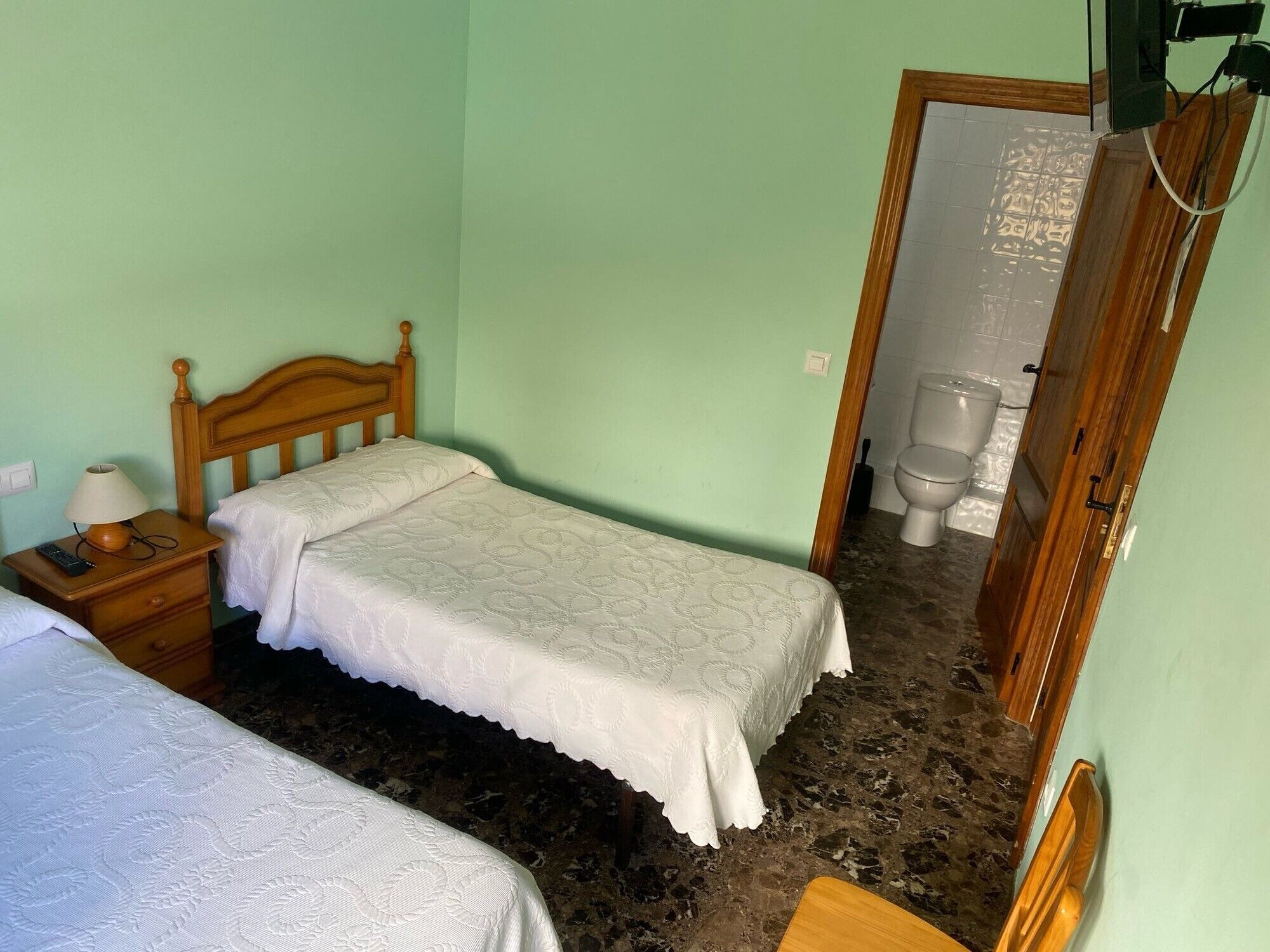 Een kleine kamer met een enkel bed, een houten hoofdbord, een nachtkastje met lamp en een zichtbare badkamer door een geopende deur.