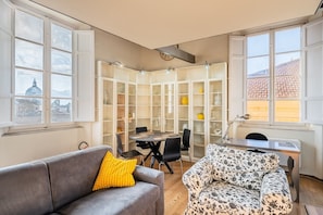 A living room with a sofa, a patterned armchair, a dining table with chairs, and a bookshelf.