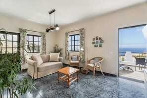 A living room with a sofa, a wooden coffee table, wicker chairs, a potted plant, and a view of the sea through a large window.