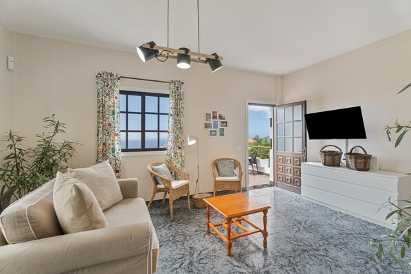 A living room with a sofa, chairs, a coffee table, a TV, and a balcony with plants.