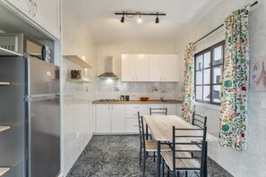 A compact kitchen with white cabinets, a stainless steel refrigerator, a wooden dining table, and a window with floral curtains.