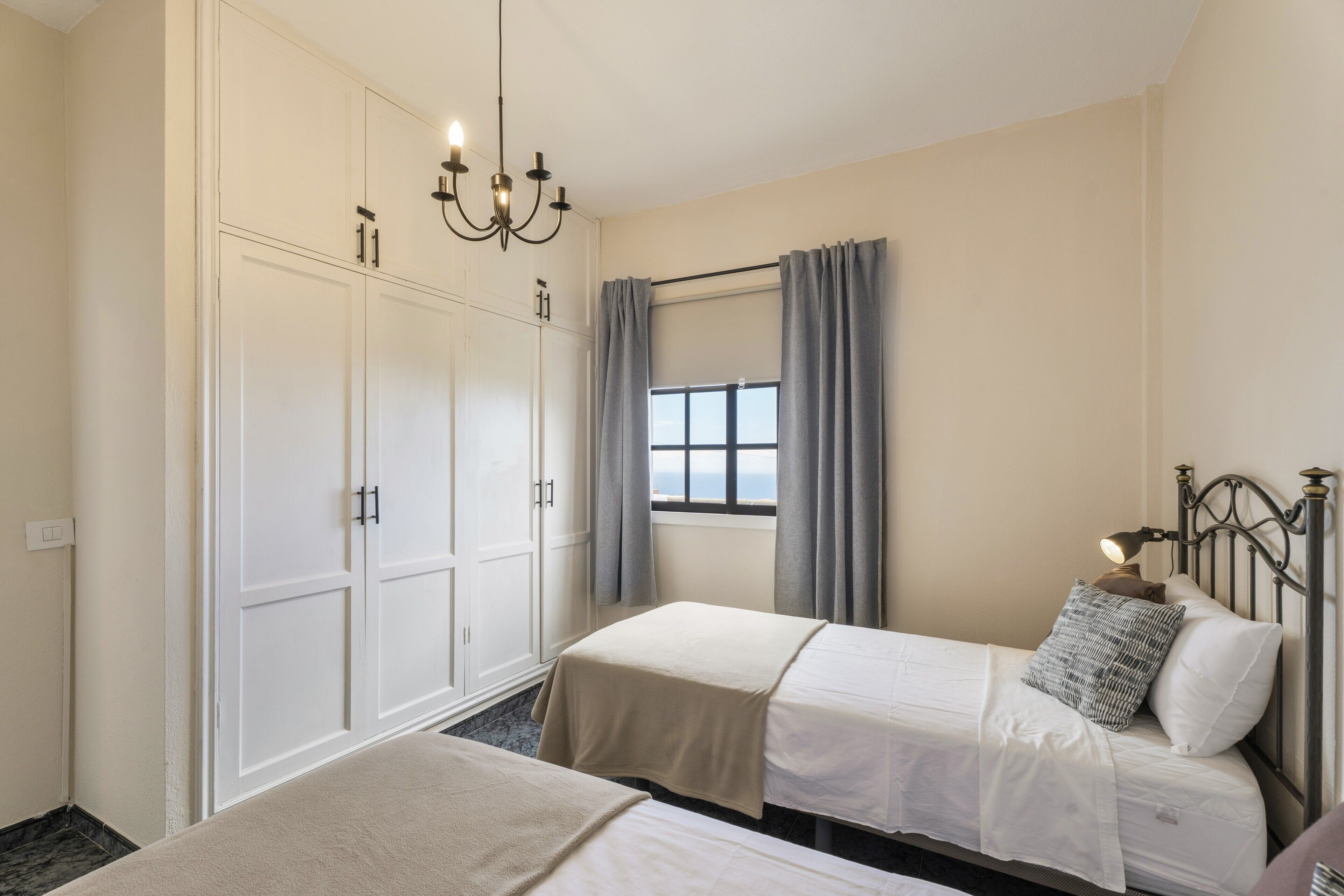 A bedroom with two beds, a wardrobe, a window with curtains, and a chandelier.