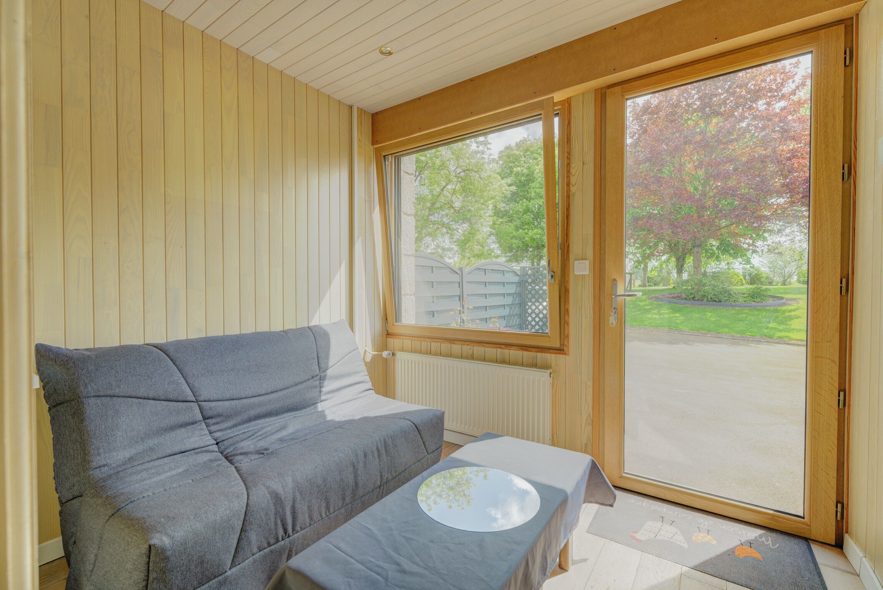 A room with a sofa, a small table, a window with a view of trees, and a door.