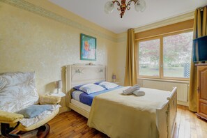 Un dormitorio con cama, una silla, un cuadro, una ventana y un televisor.