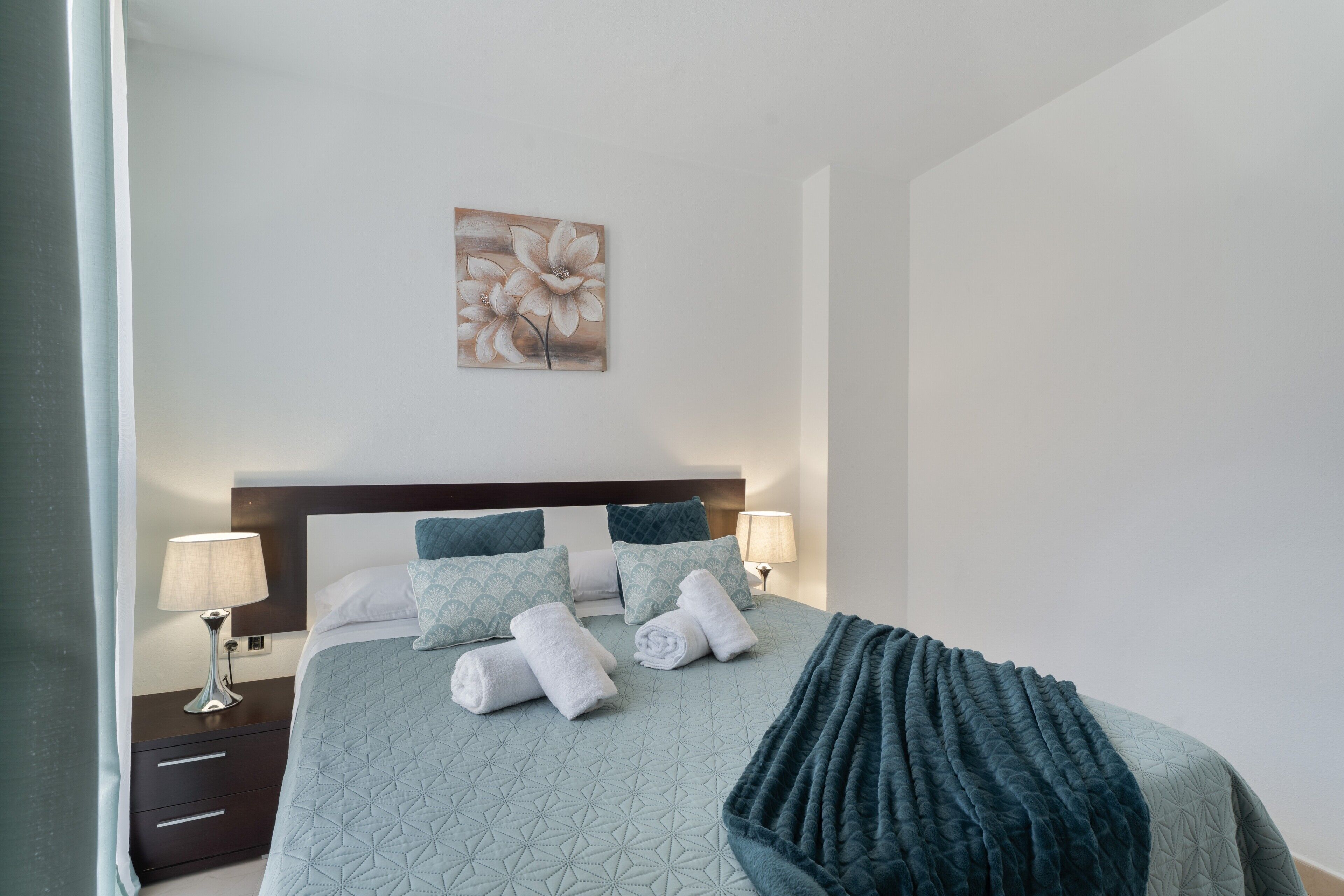 A neatly made bed with pillows and a blanket, a bedside table with a lamp, and a wall-mounted artwork.
