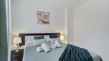 A neatly made bed with pillows and a blanket, a bedside table with a lamp, and a wall-mounted artwork.