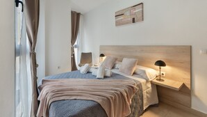 Un dormitorio con cama, almohadas, una mesita de noche con lámpara, una obra de arte en la pared y una ventana con cortinas.