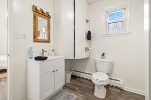 Un baño con lavamanos blanco, espejo y inodoro. Se ve una ventana con vistas a árboles y un perchero.