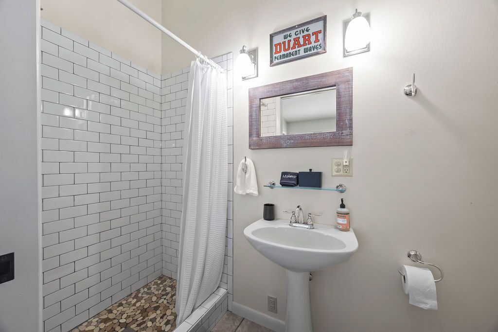 Un baño con paredes de azulejos blancos, una ducha con cortina, un lavamanos con espejo y un cartel sobre el espejo.