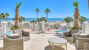 A beachside patio with wicker furniture, a pool, and palm trees.