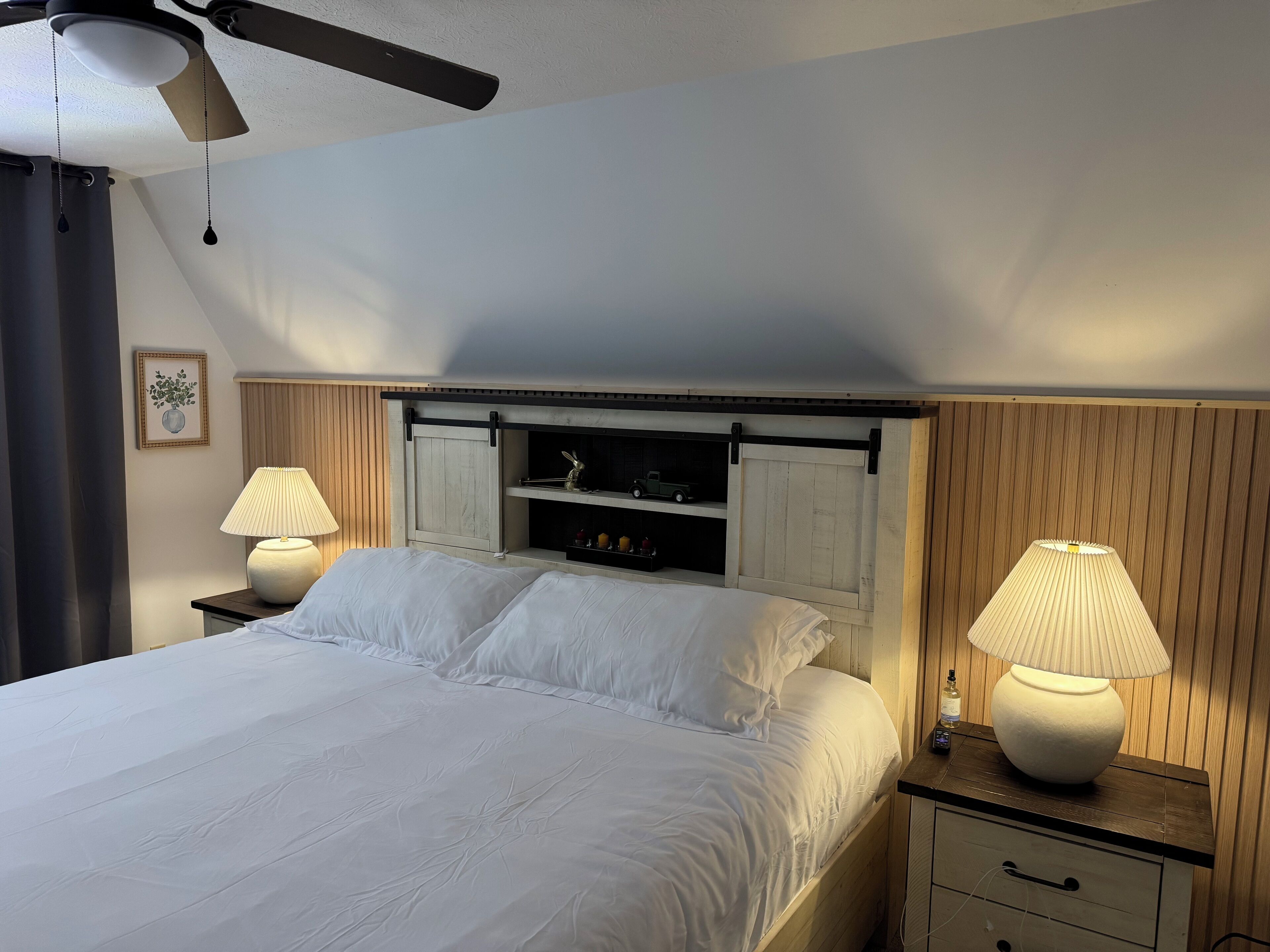 Una cama bien hecha con sábanas blancas, dos lámparas de noche, un ventilador de techo y un cabecero de madera.