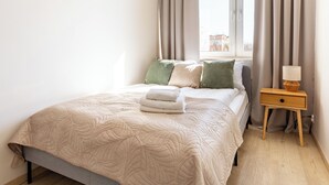 A neatly made bed with a beige quilt, a bedside table with a lamp, and a window with curtains.