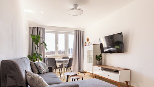 A modern living room with a grey sofa, a dining table with chairs, a TV on the wall, and a large window with curtains.