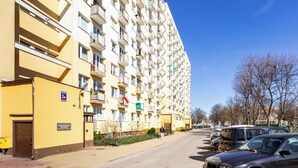 A multi-story residential building with balconies, a paved street, parked cars, and leafless trees.