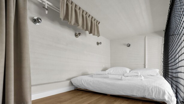 A bedroom with a wooden floor, a bed with white linens, a window with blinds, and a curtain.