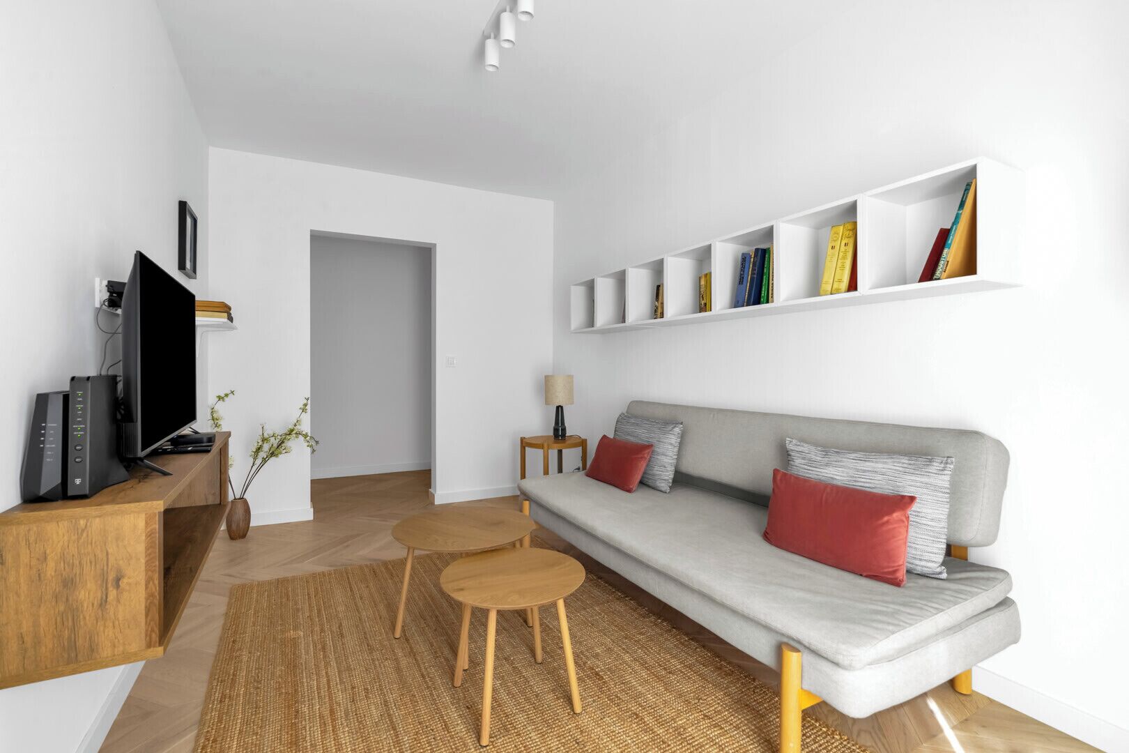 A modern living room with a grey sofa, red and grey cushions, a wooden coffee table, a TV on a wooden stand, and a bookshelf with books.