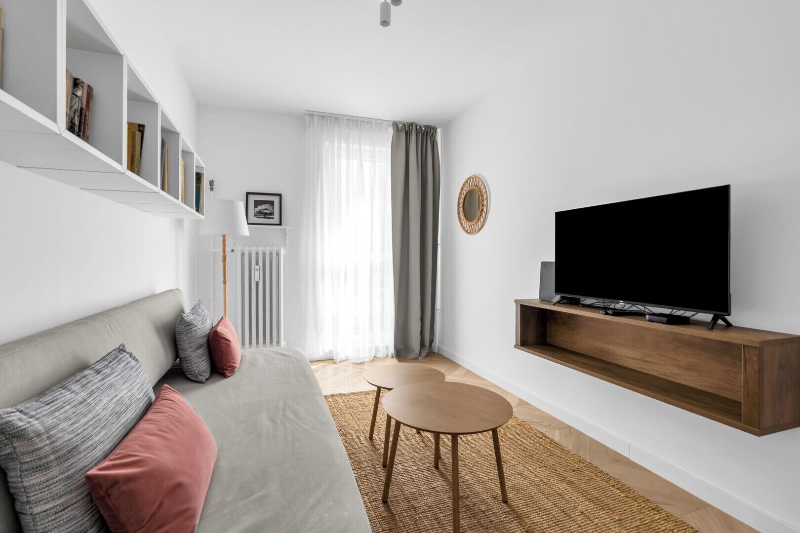 Un salon moderne avec un canapé gris, une petite table ronde, un téléviseur fixé au mur et une bibliothèque.