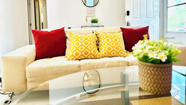 A living room with a beige sofa, red and yellow patterned cushions, a round mirror on the wall, and a potted plant on a glass table. - Cozy and Charming House (Pineville)