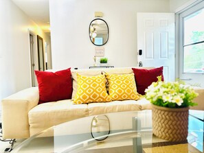 Un salon avec un canapé beige, des coussins aux motifs rouges et jaunes, un miroir rond accroché au mur et une plante en pot posée sur une table en verre.