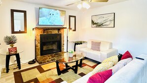 A living room with a stone fireplace, a flat-screen TV mounted above it, a ceiling fan, a white sofa with colorful cushions, and a coffee table. - Cozy and Charming House (Pineville)