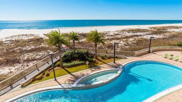 Una piscina de borde curvo, rodeada por una terraza y palmeras, con vista a una playa con dunas de arena.