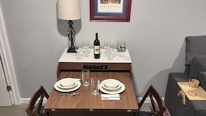 A dining area with a wooden table set for two, a framed picture on the wall, and a lamp beside the table.