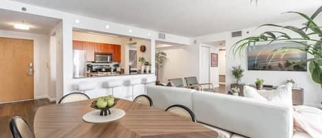 A modern living room with a dining table, white sofa, and a kitchen area in the background.