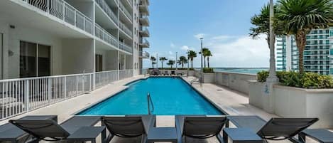 A rooftop pool with lounge chairs, a view of the ocean, and a high-rise building with balconies.