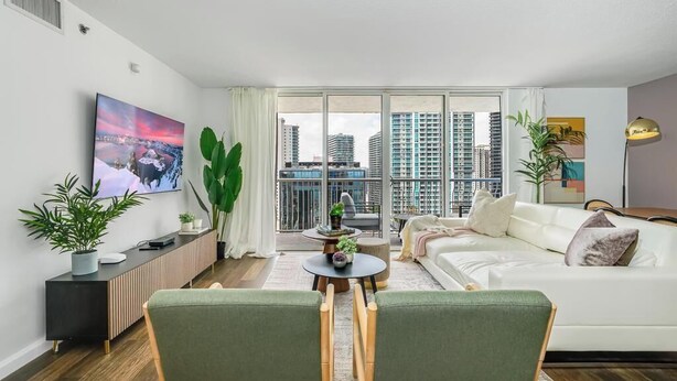 Una sala de estar moderna con un sofá blanco, un sillón verde, una mesa de centro y un televisor montado en la pared. Hay plantas sobre la mesita auxiliar y se ve una vista de edificios altos a través del amplio ventanal.