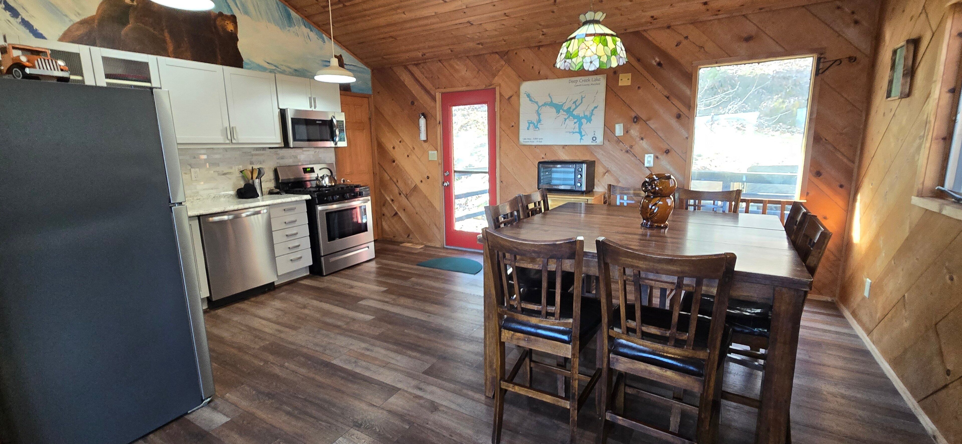 A cabin-style kitchen with wooden walls, a dining area with wooden chairs, and a large window.