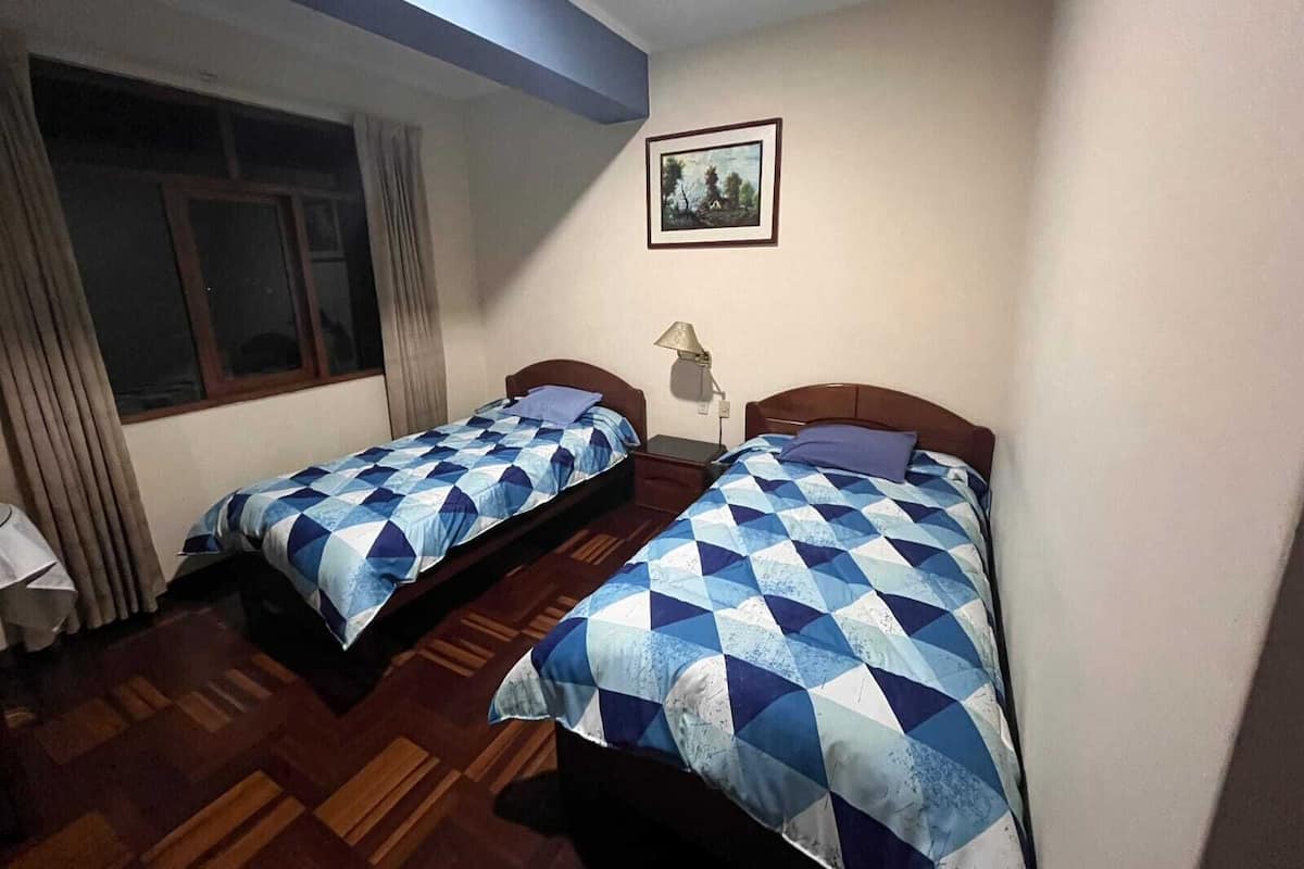 A room with two beds, a window with curtains, a lamp, and a framed picture on the wall.