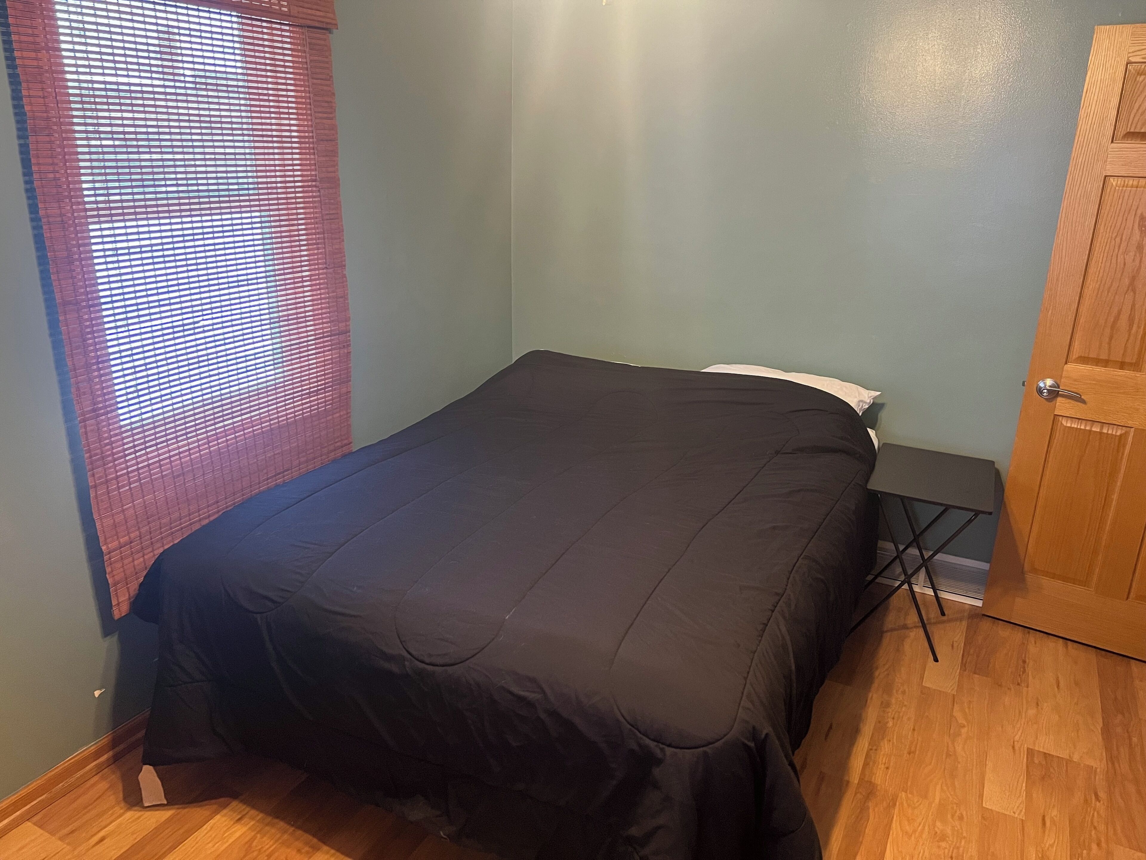 Une petite chambre avec un lit simple, un plancher en bois, une fenêtre avec des stores, une table de chevet et une porte.