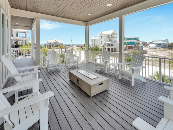 Une terrasse extérieure couverte, dotée de chaises Adirondack blanches, d’une table en béton et offrant une vue sur la plage et l’océan.