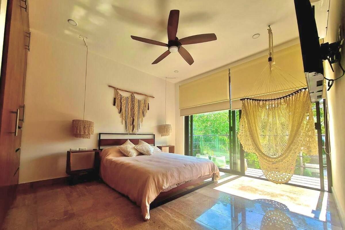 Ein geräumiges Schlafzimmer mit einem großen Bett, einem Deckenventilator, einem Fenster mit Blick auf Grünflächen und einer Hängematte am Fenster.
