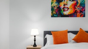 A bedroom with a bed, pillows, a nightstand, a lamp, and a colorful painting on the wall. - Stylish Brick Lane Apartment with Balcony Sleeps 4 (Greater London)