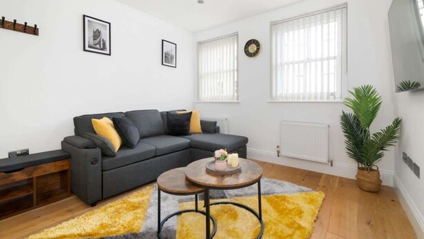 A modern living room with a sectional sofa, a coffee table, and a large window with blinds. - Stylish Brick Lane Apartment with Balcony Sleeps 4 (Greater London)