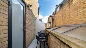 A narrow outdoor corridor with a wooden floor, a metal chair, and a glass door on one side. - Stylish Brick Lane Apartment with Balcony Sleeps 4 (Greater London)