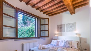 A bedroom with a large window, wooden ceiling beams, a bed with floral bedding, and two wall-mounted lamps. - Nice apartment for 4 guests with pool, A/C, WIFI, TV, terrace and panoramic view (Riparbella)