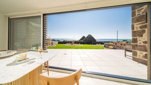 Een eetruimte met een tafel en stoelen, een glazen deur die uitkomt op een terras met uitzicht op een stenen gebouw en de zee.